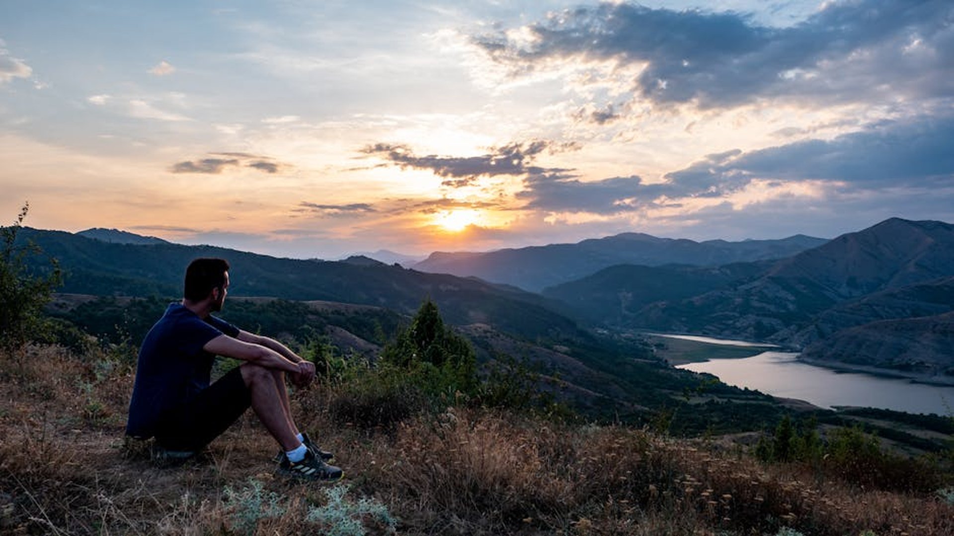 a traveler sitting on a mountain trail at sunset, backpack by their side, looking at the horizon with a quiet, reflective expression, emotional and transformative travel moment