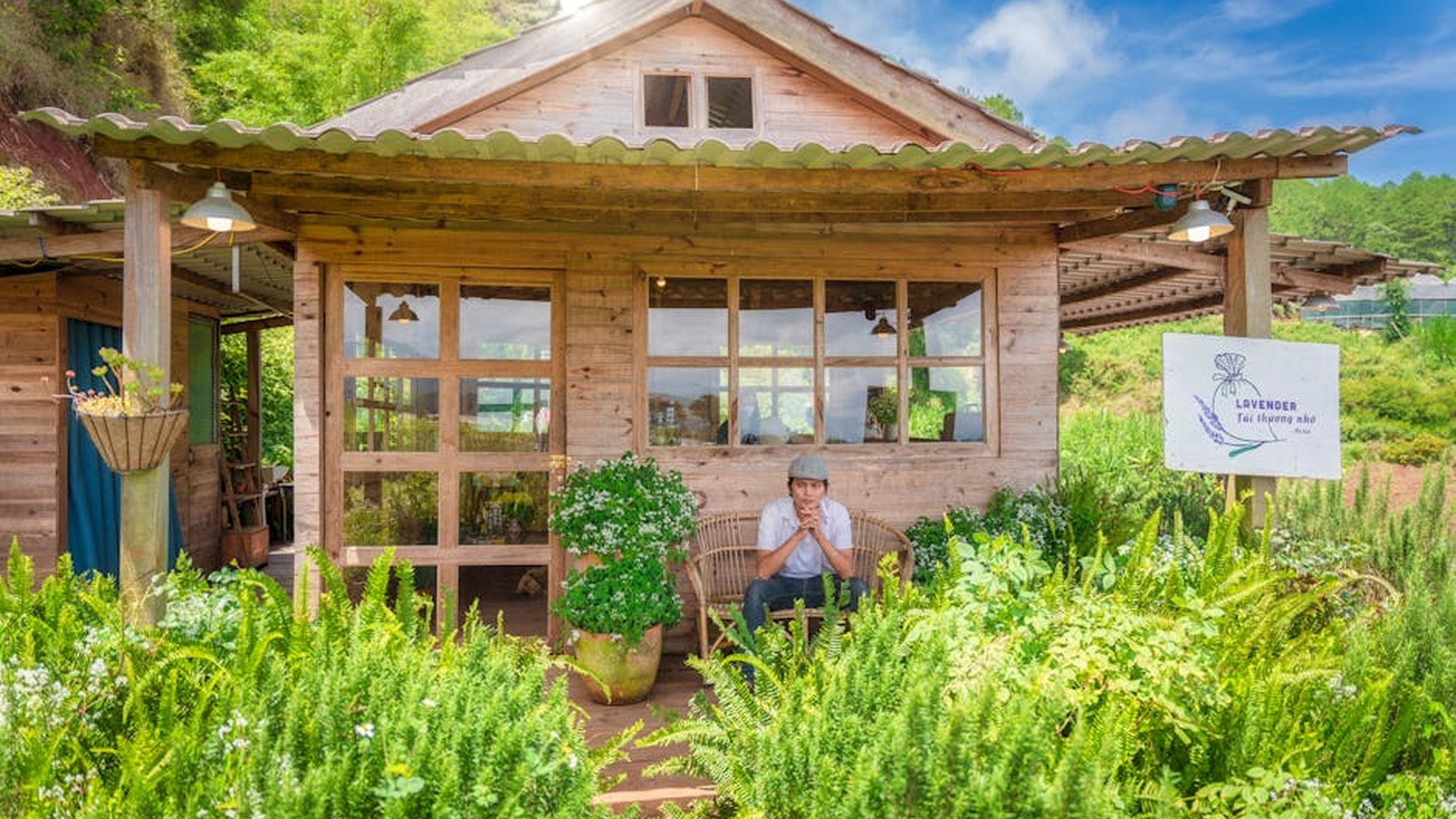a cozy homestay in a rural village, wooden cabin with a small porch, a traveler sitting outside sipping tea, surrounded by green hills and local gardens, peaceful and intimate